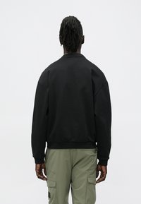 Άνδρας με dreadlocks που φοράει μακρυμάνικο μαύρο πουκάμισο και χακί παντελόνι cargo, στέκεται αντιμέτωπος σε απλό λευκό φόντο.