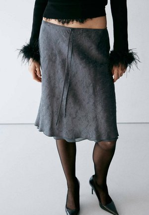Le mannequin porte une jupe grise à hauteur du genou avec une couture sur le devant, des collants noirs transparents, des escarpins pointus et un haut noir à poignets ornés de plumes.