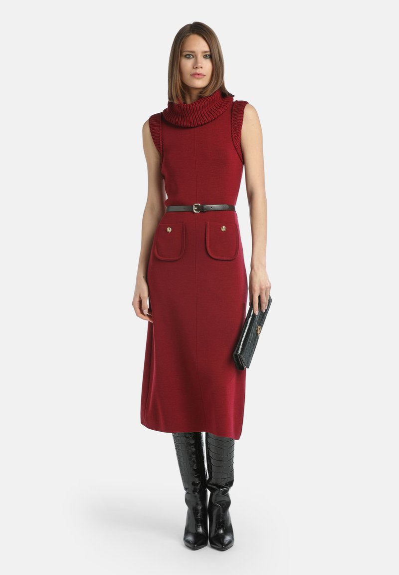 Ženska nosi brezrokavno rdečo midi obleko s turtleneck izrezom, črn pas, črne škornje do kolen in drži črno torbico.