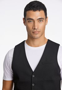 Schwarze, maßgeschneiderte Weste mit glatter Textur, ausgestattet mit einem V-Ausschnitt, fünf Knöpfen und einer Brusttasche, getragen über einem weißen T-Shirt.