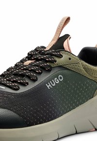 Gros plan sur une chaussure de sport noire et vert olive avec des lacets noirs à motif, une languette beige et le logo "HUGO" sur le côté.