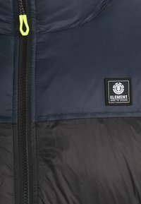 Marineblauwe en zwarte gewatteerde jas met ritssluiting, voorzien van een felgele ritstrekkertje en een zwart logo-patch op de linkerborst.