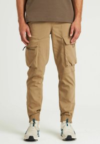 Tan cargobroek met elastische boorden, meerdere voorkorrels, een gladde textuur en een regular fit. Draagt met lichte sneakers.