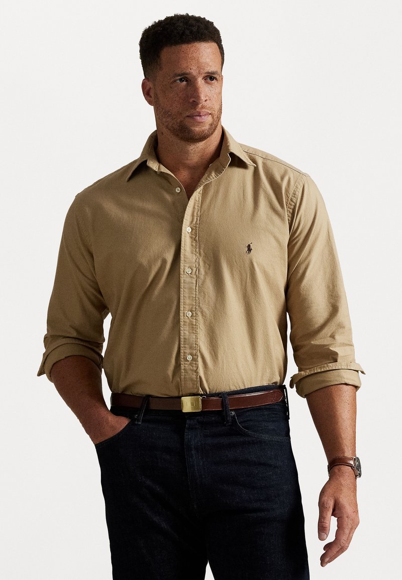 Beige gombos ing, felgöngyölt ujjal, kis hímzett logóval. Sötét farmernadrággal és barna övvel viselve.