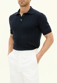 Marinblå kortärmad polotröja med en texturerad kabelstickad design, utrustad med en klassisk krage och treknapps knäppning, som är parat med vita byxor.