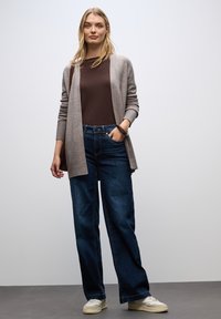Jasnoszara kardigan na brązowej bluzce, w połączeniu z ciemnoniebieskimi szerokimi spodniami dżinsowymi i beżowymi trampkami, stojąca na jednolitym tle.