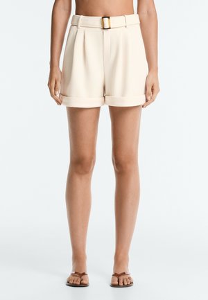 Kvinde iført beige shorts med høj talje med bælte og opkørte manchetter, kombineret med brune flade sandaler, stående mod en hvid baggrund.