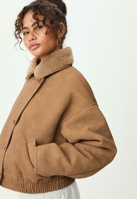 Hnědý umělý zamíkový kabát s beránkovým límcem, oversized střih, patentky a elastický pas; má boční kapsy a manžety na rukávech.