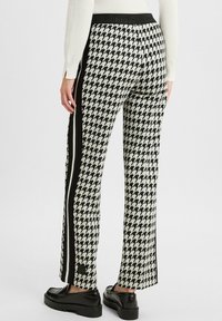 Woman wearing black and white houndstooth patterned pants with a black side stripe and black loafers, standing against a plain background.