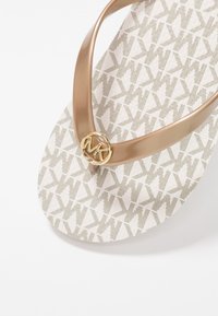 Lichtbruine flip-flop met een gouden merkaccent, voorzien van een gestippelde witte zool met kleine logomotieven en gladde banden.