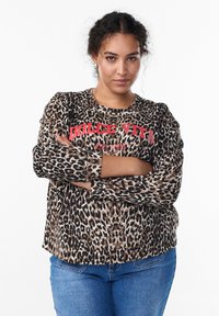 Mladá žena s kučeravými vlasmi má na sebe mikinu s leopardím vzorom a červeným nápisom „Dolce Vita“, kríži si ruky a stojí pred bielym pozadím.