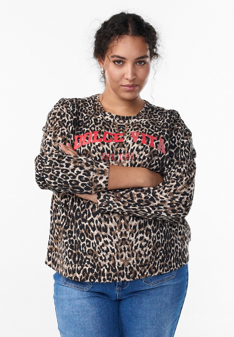 Mladá žena s kučeravými vlasmi má na sebe mikinu s leopardím vzorom a červeným nápisom „Dolce Vita“, kríži si ruky a stojí pred bielym pozadím.