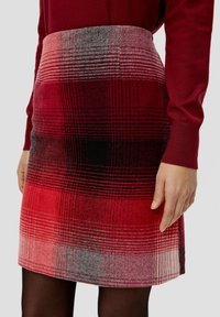 Femme portant une jupe écossaise rouge et un haut à manches longues rouge assorti, avec des collants noirs visibles sous l'ourlet de la jupe.