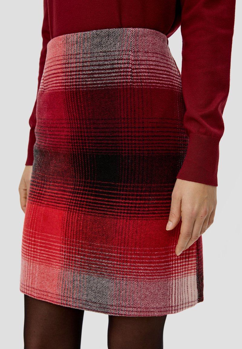Femme portant une jupe écossaise rouge et un haut à manches longues rouge assorti, avec des collants noirs visibles sous l'ourlet de la jupe.