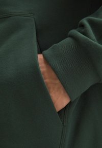 Donkergroene sweatshirt met geribbelde manchetten en een kraag, met een gedeeltelijk zichtbare hand in een zijzak die de zachte stofstructuur laat zien.