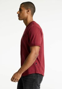 Burgunderrotes Kurzarm-T-Shirt aus Baumwolle, hat einen runden Halsausschnitt und ein kleines Logo-Detail auf der Brust. Wird mit dunklen Jeans getragen.