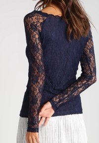 Haut en dentelle bleu marine à manches longues avec motif floral, manches transparentes et corps uni. Associé à une jupe argentée à franges.