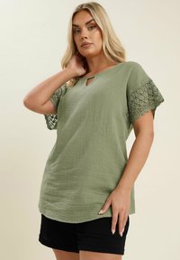 Olivgrön blus med en texturerad yta, med ett nyckelhålsutskärning och korta ärmar med spetskant. Matchad med svarta shorts.