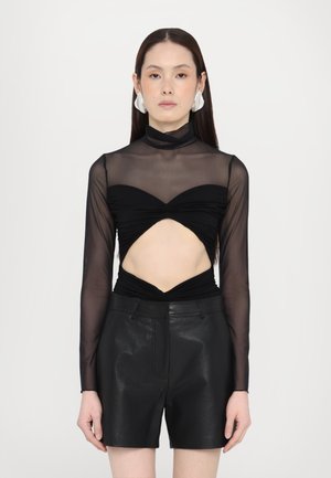Sort langærmet top med gennemsigtig mesh-stof, der har et udskæringsdesign i taljen. Kombineret med sorte lædershorts og markante øreringe.