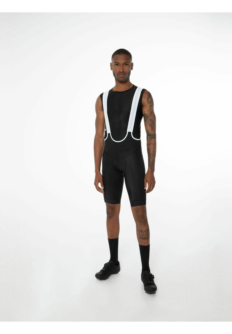 Schwarzes Radoutfit mit weißen Trägern, aus atmungsaktivem Material. Enganliegendes Design, kurze Beine und sichtbare, knöchellange schwarze Socken.