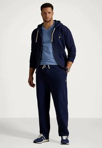 Άνδρας ντυμένος με μπλε φούτερ ζακέτα με φερμουάρ, αντίστοιχες φόρμες, ανοιχτό μπλε μπλουζάκι και μπλε αθλητικά παπούτσια με λευκές λεπτομέρειες.