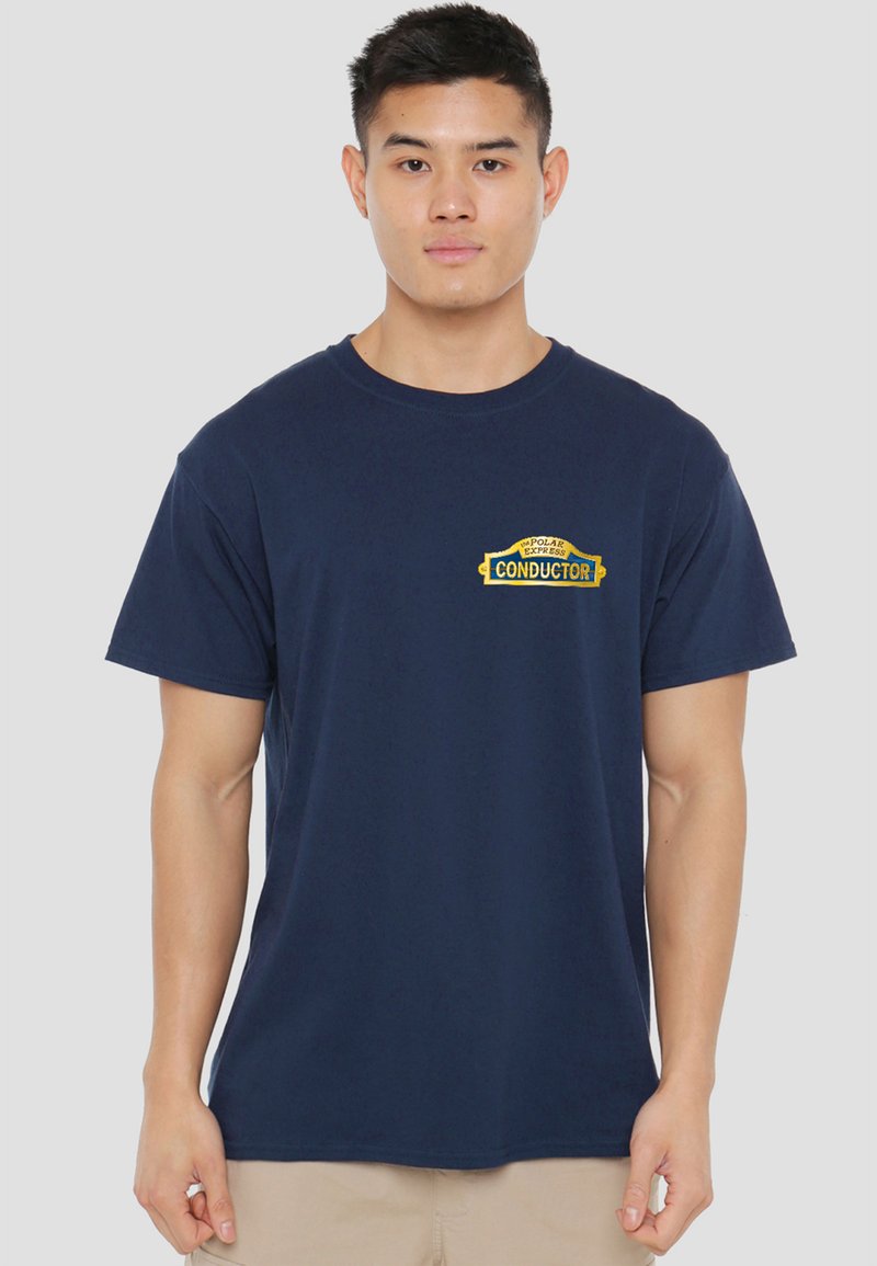 Young man standing, wearing a navy blue t-shirt with a yellow "Conductor" patch on the left chest and beige pants.