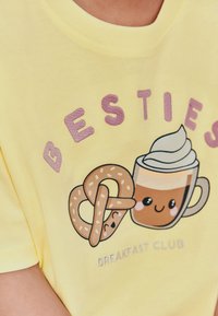 Kollane lühikeste varrukatega T-särk, millel on mänguline kujutis naeratavast kohvitassist ja pretzlist ning lilla tekstiga "BESTIES" ja "BREAKFAST CLUB".