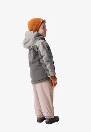 Kind trägt eine graue und olivgrüne Jacke mit Kapuze, beige Hosen und orangefarbene Handschuhe, steht seitlich vor einem weißen Hintergrund.