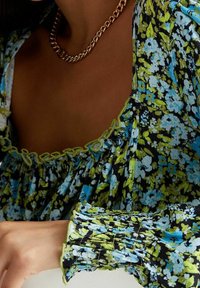 Blommig blus i svart med blå och gröna mönster. Har en samlad halsringning och puffärmar med kontrasterande kant. Guldhalsband.