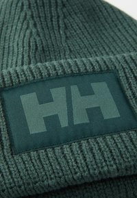 Strikket lue i mørkegrønn med ribbet tekstur. Har en rektangulær logo-lapp som viser "HH" i en lysere nyanse av grønn.