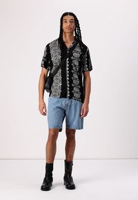 Cămașă neagră cu mânecă scurtă și modele florale albe, asociată cu pantaloni scurți din denim albastri și cizme negre la gleznă; geantă mică neagră tip crossbody.