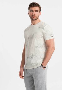 Camiseta de manga corta en color beige claro con motas grises dispersas, diseño de cuello redondo y un pequeño detalle de logo en la manga izquierda.