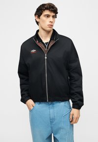 Νέος άνδρας που φοράει μαύρο μπουφάν Umbro με φερμουάρ πάνω από αθλητικό μαύρο τοπ και ανοιχτό μπλε τζιν με ξεφτισμένες λεπτομέρειες, με τα χέρια στις τσέπες, κοιτάζοντας δεξιά.