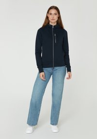 Sudadera con cremallera color azul marino, con capucha, que presenta una cremallera en el frente, bolsillos y puños acanalados. Combinada con jeans de color azul claro y zapatillas blancas.