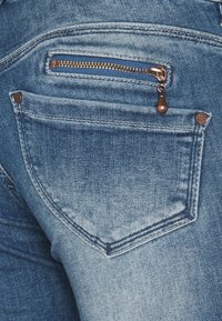 Zadní kapsa džín s měděným zipem a bronzovým nýtem jako ozdobou. Tkanina je světle modrá s jemným vyblednutím a hladkou texturou.