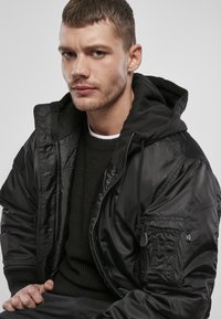 Giacca bomber nera con texture lucida e trapuntata; presenta un cappuccio, zip frontale e tasche laterali. Indossata sopra un maglione nero in maglia.