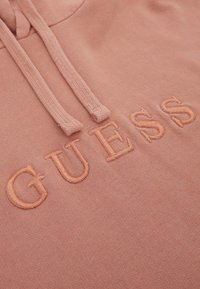 Sudadera rosa claro con un acabado texturizado, cordones ajustables y el logotipo bordado "GUESS" en hilo a juego.