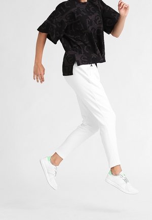 Sort oversized t-shirt med fed, tekstureret skrift, hvide tilspidsede bukser og hvide sneakers med grønne detaljer, i et springende positur.