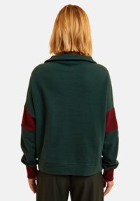 Felpa verde con maniche lunghe, dettagli bordeaux sui polsi e sulle spalle, tessuto morbido, vestibilità rilassata e colletto a risvolto.
