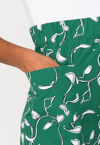 Tissu vert avec des motifs floraux blancs, doté d'une taille élastique et de poches latérales. Le design comprend un motif de feuille entouré de noir.