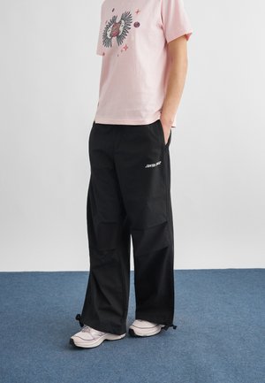 Osoba oblečená v ružovom tričku s potlačou, voľných čiernych nohaviciach Santa Cruz a bielych teniskách stojaca na modrom koberci s rukami v vreckách.