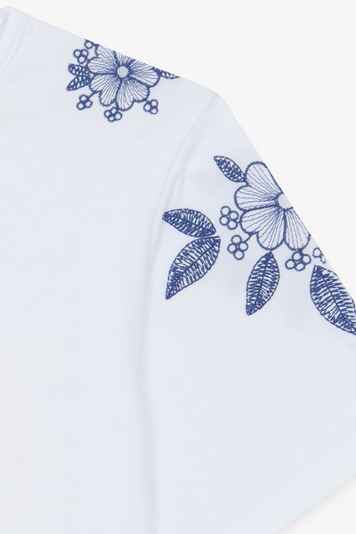 Λευκό βαμβακερό μπλουζάκι με μπλε κεντήματα λουλουδιών στο μανίκι, που περιλαμβάνουν λεπτομερείς σχεδιασμούς φύλλων και λουλουδιών. Απαλή υφή, άνετη εφαρμογή.