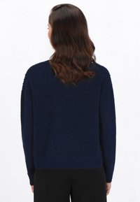 Vrouw met schouderlang bruin haar, gekleed in een marineblauwe lange mouw sweater en zwarte broek, van achter afgebeeld tegen een witte achtergrond.