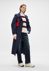 Trench coat navy peste un pulover alb cu dungi roșii, asortat cu blugi de culoare albastră închisă și teniși de culoare deschisă. O geantă roșie adaugă detaliu.