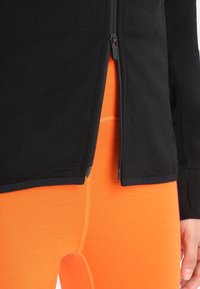 Giacca nera con cerniera parzialmente aperta sopra pantaloni aderenti di colore arancione brillante, che mostrano la trama dettagliata del tessuto e le cuciture.