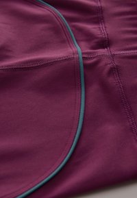 Tessuto atletico viola con una texture liscia, caratterizzato da piping turchese a contrasto lungo le cuciture e i contorni, che evidenzia un design aderente.