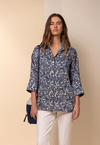 Blusa a fantasia paisley blu e bianca con maniche a tre quarti, apertura con bottoni, abbinata a pantaloni bianchi e una borsa blu.