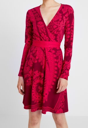 Frau trägt ein langärmeliges, rotes Wickelkleid mit dunkelrosa Blumen- und Paisley-Mustern und einem passenden Taillengürtel.