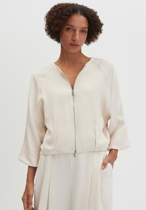 Vrouw met kort krullend haar die een crème kleurige lange mouwen jas met rits en bijpassende crème kleurige broek draagt, staand met één hand in de zak.