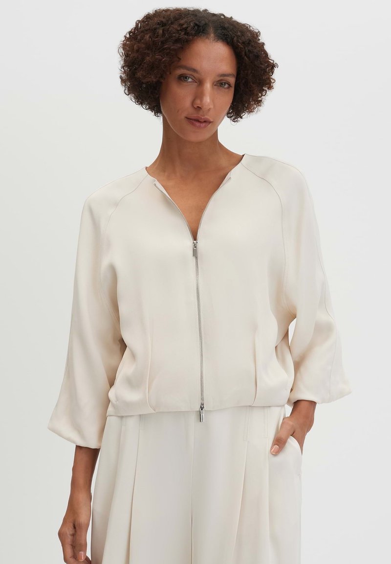 Vrouw met kort krullend haar die een crème kleurige lange mouwen jas met rits en bijpassende crème kleurige broek draagt, staand met één hand in de zak.